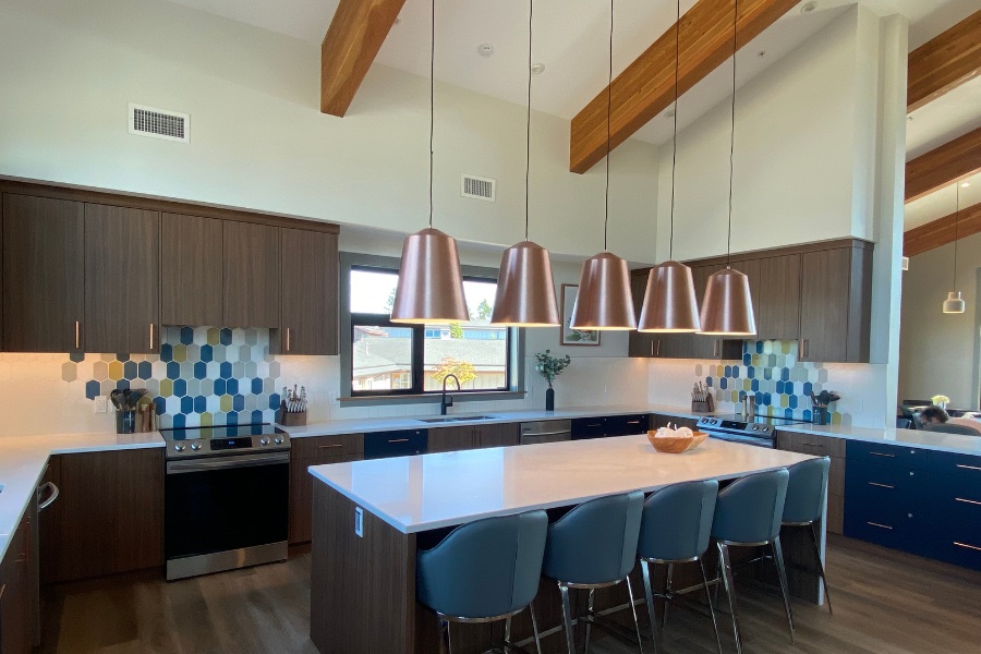 A kitchen with dark wood cabinets, beams on the angled ceiling, white countertops, and blue stools at the island, located in the Qwalayu House, designed by Alan Lowe Architecture.