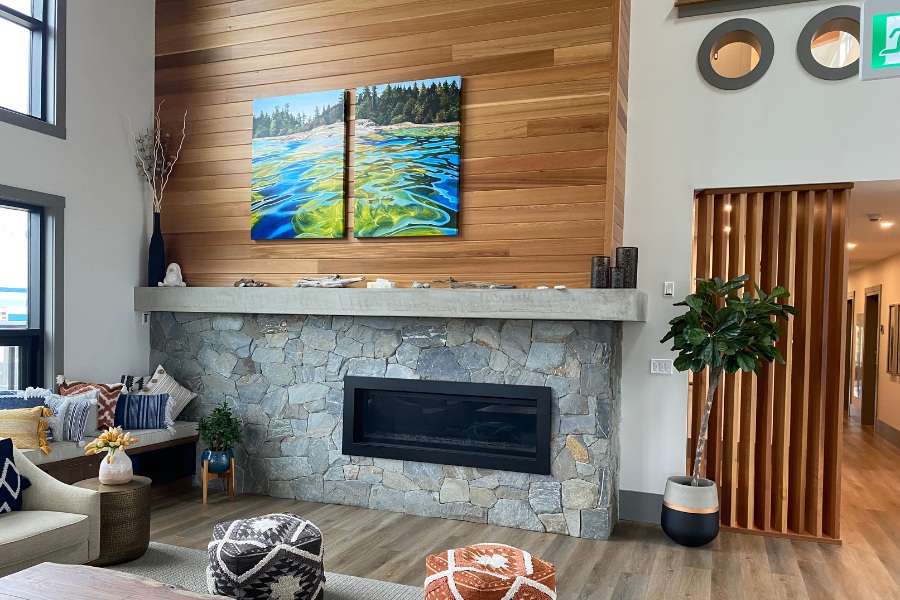 A living room with large windows, a stone fireplace with accents of wood, in the Qwalayu House, designed by Alan Lowe Architecture.