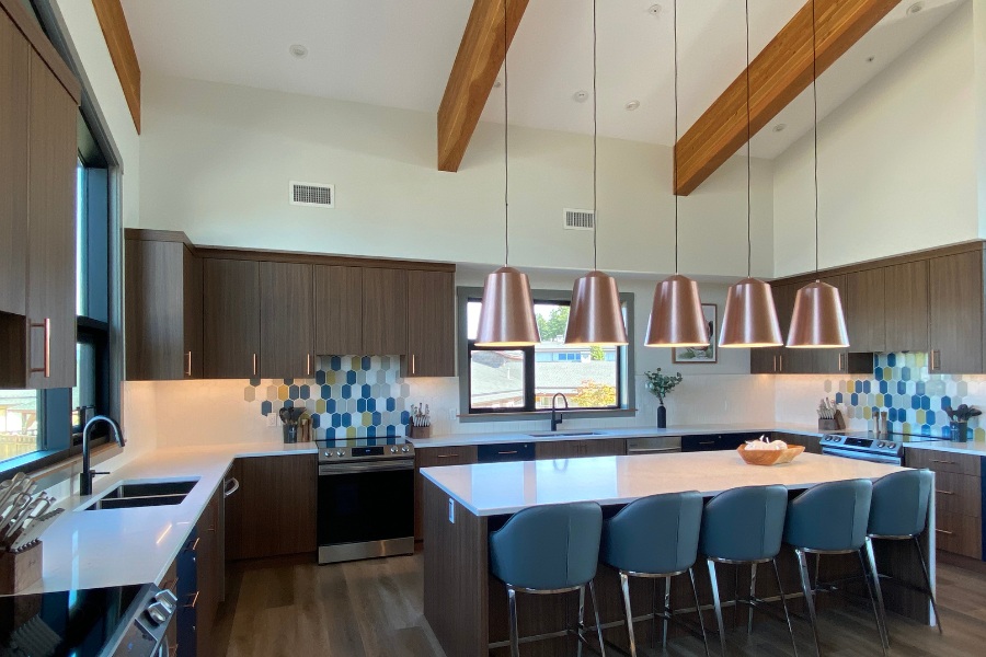 A kitchen with dark wood cabinets, beams on the angled ceiling, white countertops, and blue stools at the island, located in the Qwalayu House, designed by Alan Lowe Architecture.