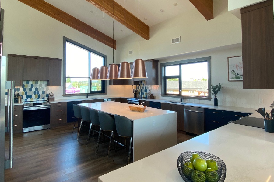 A kitchen with dark wood cabinets, beams on the angled ceiling, white countertops, and blue stools at the island, located in the Qwalayu House, designed by Alan Lowe Architecture.