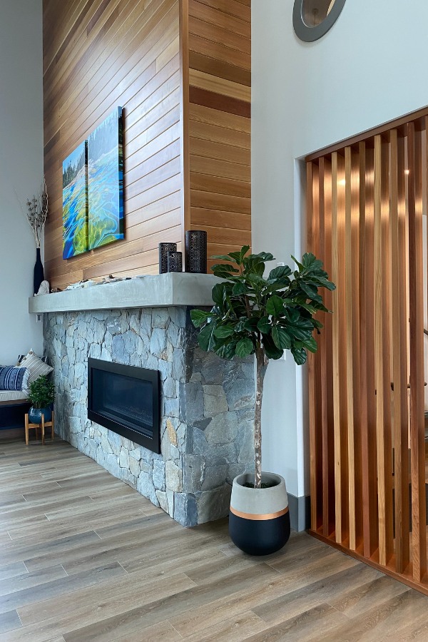 A living room with large windows, a stone fireplace with accents of wood, in the Qwalayu House, designed by Alan Lowe Architecture.