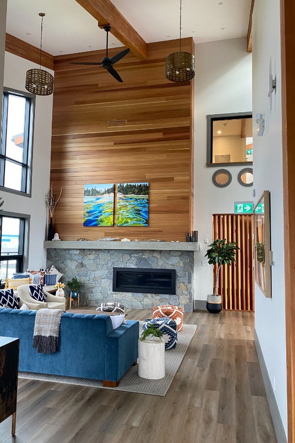 A living room with large windows, a stone fireplace with accents of wood, in the Qwalayu House, designed by Alan Lowe Architecture.